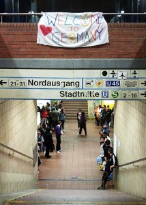 Blick von einer Bahnsteigtreppe. Am Fuß der Treppe stehen Menschen, über der Treppenbrüstung hängt ein Transparent mit der Aufschrift Welcome to Germany".