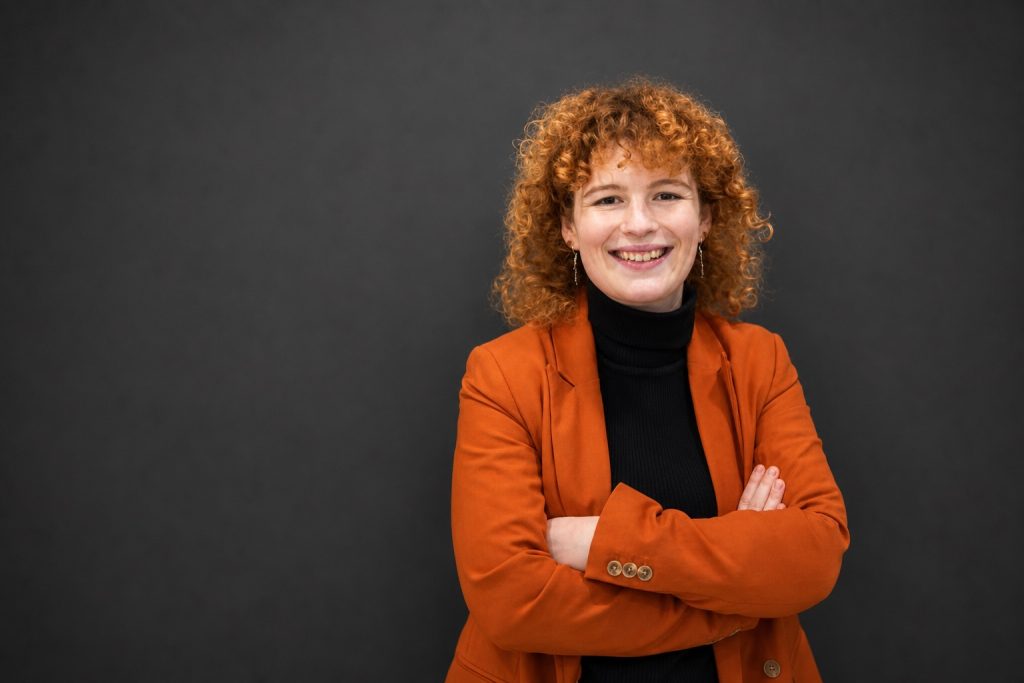 Lisa „Lizzy“ Schubert mit verschränkten Armen in orangefarbenem Blazer vor dunkelgrauem Hintergrund.