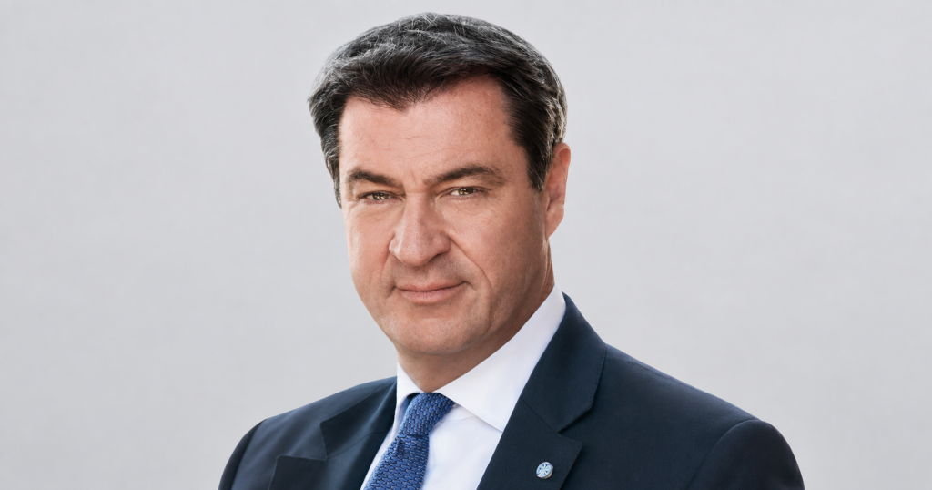 Porträt von Markus Söder vor grauem Hintergrund, seitlich mit KI zum Querformat erweitert.